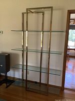Front view of the brass toned metal etagere with four clear tempered glass shelves, showing the full height and width of the piece against a wall.