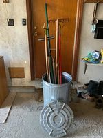 Full view of galvanized garbage can containing multiple garden tools with handles extending upward.