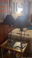 Pair of metal based lamps with black tapered shades placed on a glass top side table beside a beige couch.