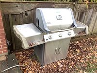 Front view of the Napoleon Prestige BBQ grill showing the stainless steel body, control knobs, side shelves, cabinet base, and natural gas hose connection.