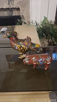 Photo showing the brass cloisonne rooster and pig figurines placed on a glass surface with fireplace and vegetation background.