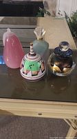 Four decorative jars on a reflective glass surface including a pink pear-shaped jar, colorful ceramic jar, dark gold-accented decanter, and green jar with flared stopper.