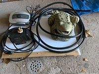 Concrete frog fountain sculpture alongside electrical pumps and black hoses laid on a board on the ground.
