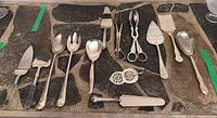 Full spread of silver plated serving utensils including various spoons, tongs, slicers, servers and utensils with decorative handles, laid out on a stone surface.