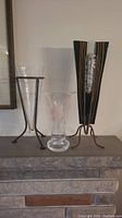 Photo showing all three vases on a mantle: tall clear glass vase on the right with metal frame, middle printed vase, and left tallest handcrafted vase