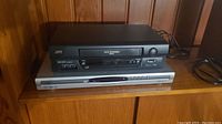 JVC VCR and Sanyo DVD player shown stacked on wooden surface with power cords visible