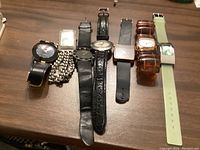 Seven assorted costume watches displayed on a wooden surface, showing variety in strap material and watch face designs.