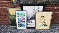 Four framed art prints leaning against a brick wall showing varied subject matter including landscapes and a portrait