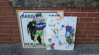 Full view of both artworks together, showing the framed Mardi Gras poster and the abstract painting leaning against a brick wall.