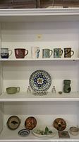 Front view of shelving holding an assortment of six hand-painted mugs, a floral plate, small bird figurines, oil burner, and various decorative pottery bowls and plates below.