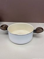 Front angled view of white Moneta ceramic pot showing round shape, cream inner ceramic coating, and two brown handles with vented design.