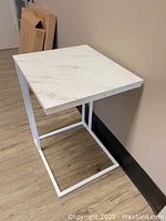 Fully assembled white faux marble side/end table with square top and white metal C-shaped frame on wood floor.