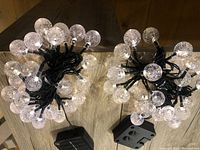 Two separate sets of solar globe string lights with bubble style LED bulbs, powered by compact solar panels, arranged on a wooden floor.
