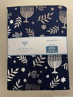 Front folded view of navy blue tablecloth with silver Hanukkah patterns and Storehouse brand label showing dimensions and product name.