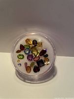 Multiple loose faceted gemstones displayed inside a clear round container filled with white padding. Variety of colors and shapes are visible.