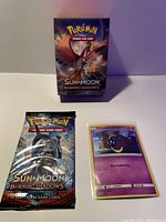 Photo showing the new empty pre-release deck box, sealed 10-card booster pack, and holographic Cosmog promo card in a protective sleeve, displayed on a flat surface with clear lighting.