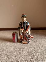 Front view of the colorful porcelain statue next to a soda can for size reference, showing detailed facial features, intricate robe design and accessories.