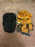 Photo showing the Deuter Rack Pack Uni black backpack side by side with the orange Arcteryx Cicero 35 backpack, displaying overall size and condition.