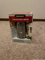 Front view of unopened Defiant keypad deadbolt retail box showing product image, satin nickel finish, and packaging details on carpeted floor.