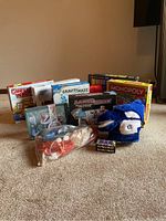 Photo depicting full view of assorted games and puzzles arranged on carpet, including Monopoly Junior, Guess Who, Laser Maze, Gravity Maze, a plush blue slipper, and Frozen themed tin.