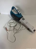 Black & Decker handheld vacuum with white and teal body placed on a charging base with attached power adapter cord.