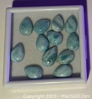 Close-up of Larimar gemstones in white square display case on purple surface. 12 polished stones in blue and white tones, teardrop and oval shapes.