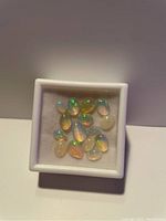Photo showing a white square container with 12 small varied cabochon Ethiopian opals exhibiting flashes of green and orange, some with light translucency.