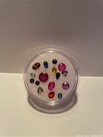 Top view of clear round container showing assortment of 18 loose sapphires in various colors and cuts placed on white cushion inside container.