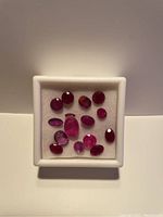 Top view of assorted ruby gemstones in a white square container showing various shades and cut styles.