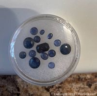 Top view of a clear round container with 16 blue sapphires of mixed cuts and sizes on white foam.