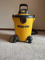 Front view of yellow Shop Vac wet/dry vacuum with black accents, showing general condition and structure including handle and wheels.
