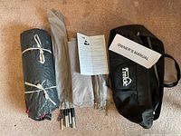 Photo showing tent components: rolled tent fabric with white rope, folded tent poles, two instruction sheets, and black carrying bag with Trekk logo.