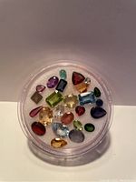 Clear round container showing multiple natural cut gemstones of various colors and cuts on white padding background