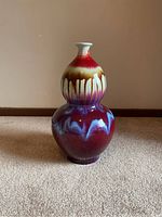 Front view of tall double-gourd shaped vase with vibrant dripped red, cream, purple, and blue glaze on carpeted floor against neutral wall.