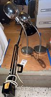 Two chrome desk lamps with spherical reflective shades, flexible gooseneck stems, and round chrome bases alongside a black articulating arm lamp with clamp base on wooden surface.