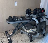 Overall view of the Bowflex adjustable dumbbells on stand, Marcy workout bench with weights on top, and foot massager beneath bench.