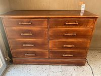 Front view of an eight drawer wood dresser with wood handles and medium brown finish placed on a tiled floor.
