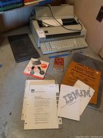 IBM Wheelwriter 5 electric typewriter with keyboard and LCD display, accompanied by printed manuals, a ribbon cassette box, a protective black cover, and various documents on a corner floor space.