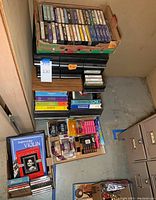 Top-down view of multiple boxes containing cassette tapes, VHS tapes and DVDs. Violin learning tapes and some audio equipment-related items visible.