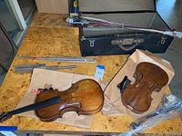Two violins with multiple bows, black trunk, and metal music stand on a table.