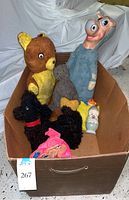 Overview of vintage stuffed animals and toys in cardboard box showing large teddy bear, poodle, gray cat, Yosemite Sam puppet, blue chick figurine, and Quick Draw McGraw doll.