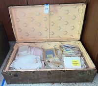 Vintage trunk open showing assortment of vintage linens, doilies, napkins, and handkerchiefs arranged inside with labels on some items