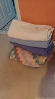 Stack of three folded bed blankets including a white knit blanket, a medium blue blanket, and a multicolored patchwork quilt on carpeted floor