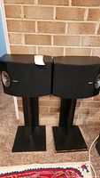 Pair of black Bose Model 301 V floor standing speakers on black stands in front of a brick wall.