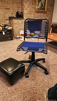 Adjustable desk chair with black metal frame and blue elastic seat and backrest beside black textured footstool with metal trim on carpet.