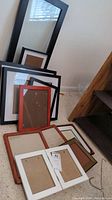 Assorted picture frames stacked and arranged on carpeted floor, sizes ranging from small to large, mix of wood and metal frames including white, brown, and black finishes.