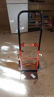 Full front view of 4 Wheel Wonder hand truck with orange frame and black handle in garage setting.