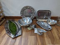 Full view of nine metal serving pieces placed on wooden floor against white wall including bowls, trays, and gravy boat showing overall shape and patina.