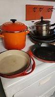 Orange enamel Le Creuset stockpot with lid, stacked black frying pans, orange enamel frying pan, and shallow baking pan on stove.