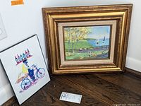 Two signed framed artworks side by side on the floor, one a colorful print and the other a framed painting on tin with a gold frame.